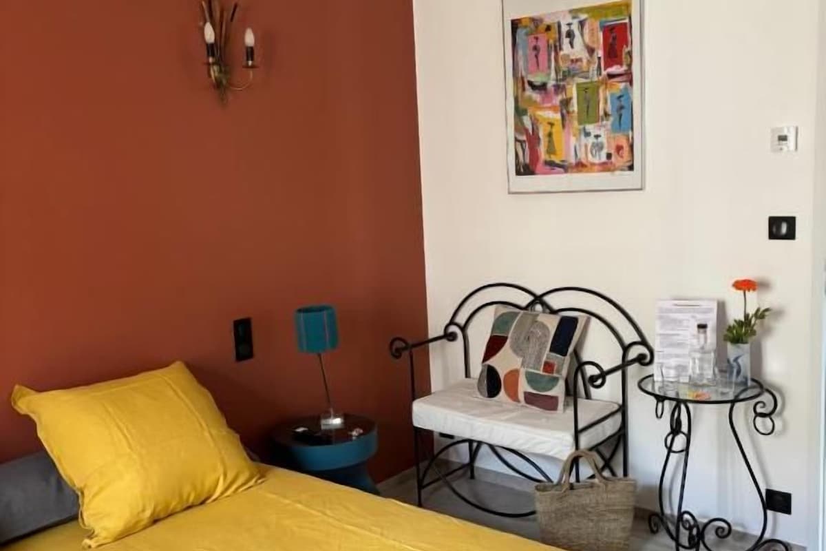 Et soveværelse med sengetæppe i gult, en stol med metalstel, et lille bord med en plante og en vægmonteret lampe.