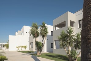 Un bâtiment moderne aux murs extérieurs blancs, doté de nombreux balcons et agrémenté d’un jardin bien entretenu avec des palmiers et de la pelouse.
