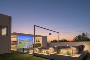 Ein moderner Außenbereich mit Sitzgelegenheiten, ausgestattet mit einem großen Bildschirm, der eine schöne Landschaft zeigt, umgeben von gut beleuchteten Pflanzen und einem zeitgemäßen Design.