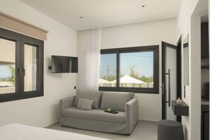 Ein modernes Schlafzimmer mit grauem Sofa, einem flachbildfernseher an der Wand und einem großen Fenster mit Blick ins Freie.