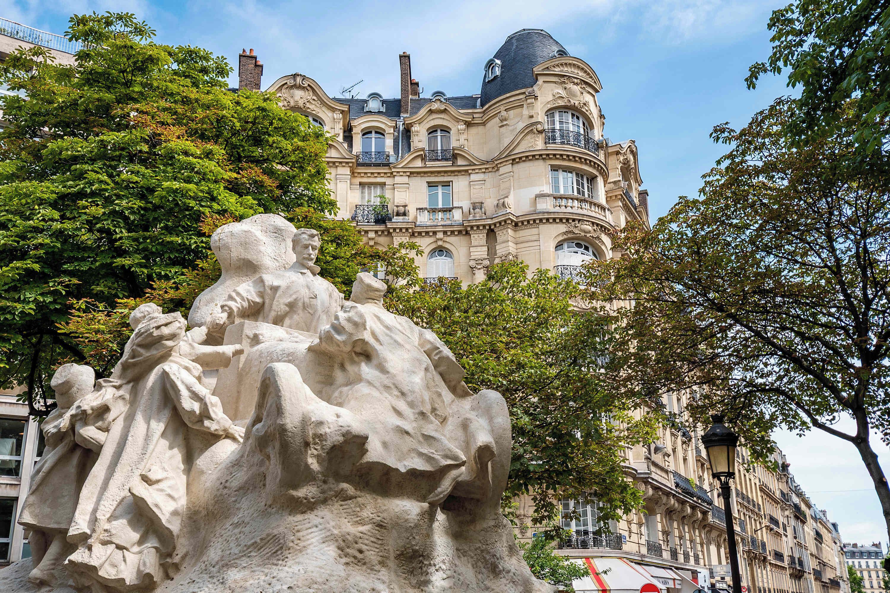 Una escultura de piedra clásica en una plaza pública, con un edificio grande y ornamentado al fondo.
