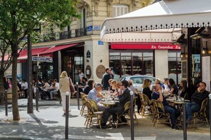 Ein Straßencafé mit Tischen und Stühlen, umgeben von Gebäuden und Bäumen.
