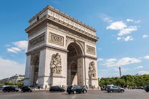 Ein großes, historisches Steinportal mit aufwendigen Schnitzereien und Inschriften, umgeben von einer belebten Straße mit Autos und Fußgängern.