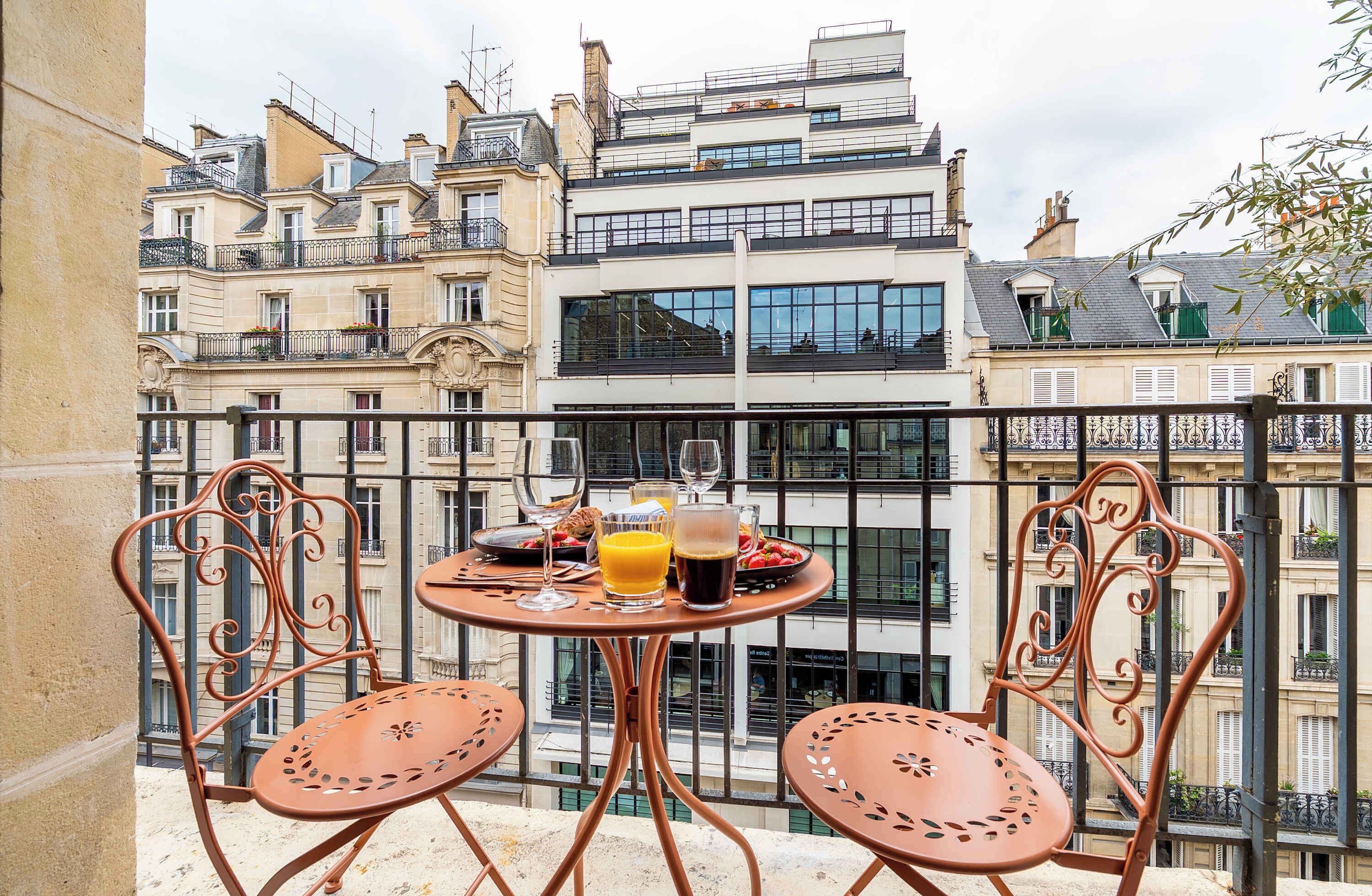 Un balcon avec deux chaises en métal et une petite table ronde, dressée pour le petit-déjeuner, avec des aliments et des boissons.