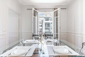 A dining area with a glass table set for two, a view of a balcony with a railing, and a white interior with paneled walls. - 2-bedroom near La Défense - Mobility Lease (Levallois-Perret)
