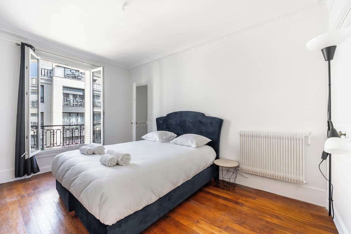 Ein Schlafzimmer mit Bett, Heizkörper, einem Fenster mit Blick auf Gebäude und einer Stehlampe.