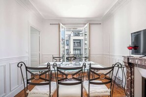 A dining area with a glass table and metal chairs, a television on the right, and a view of a multi-level building through an open door. - 2-bedroom near La Défense - Mobility Lease (Levallois-Perret)