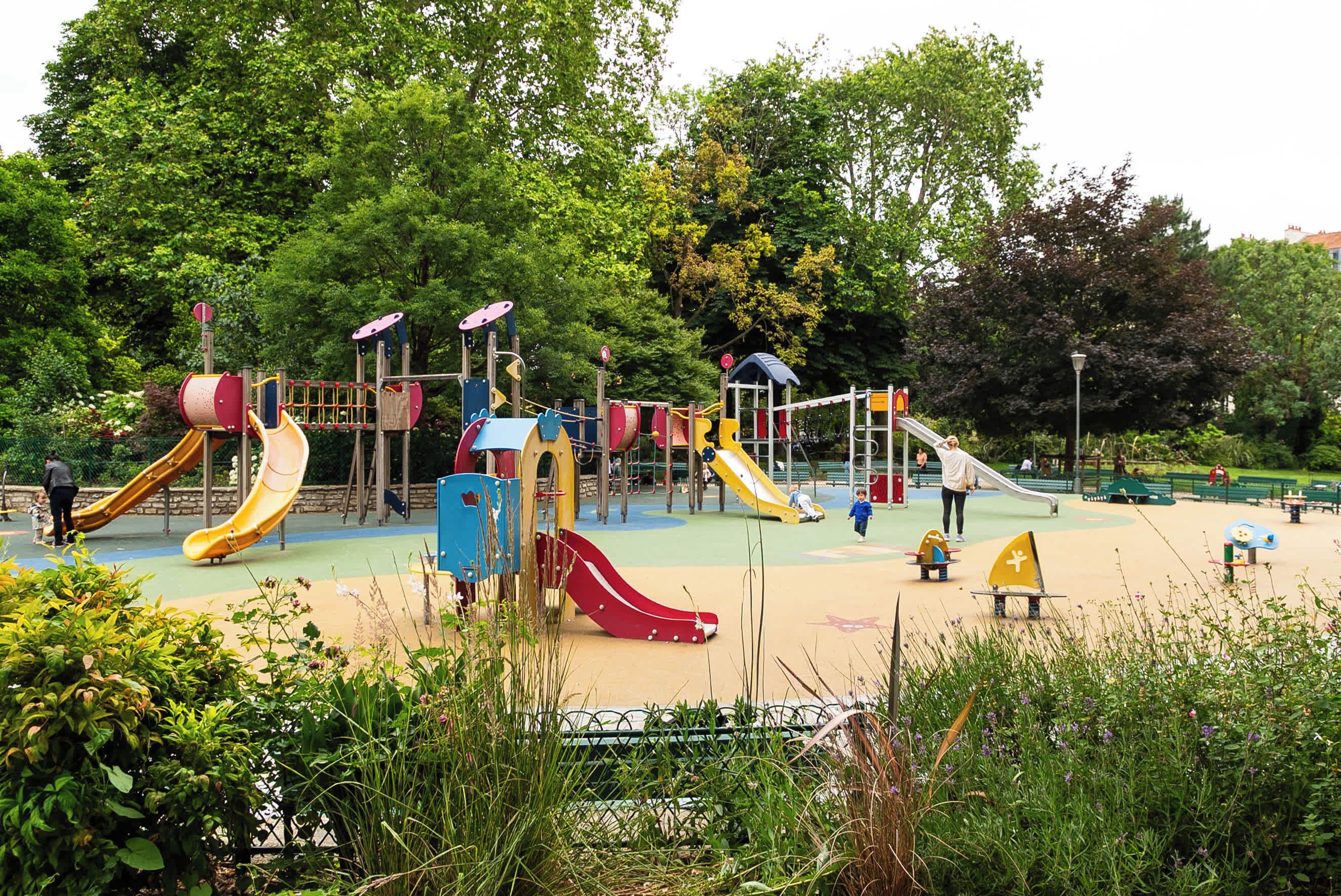 Ein Spielplatz mit verschiedenen Spielgeräten, darunter Rutschen und Kletterbereiche, umgeben von Grünflächen und Bäumen.