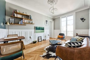A living room with a sofa, a coffee table, a television, and bookshelves. - Chic Parisian 1BR Flat near Montparnasse (Paris)