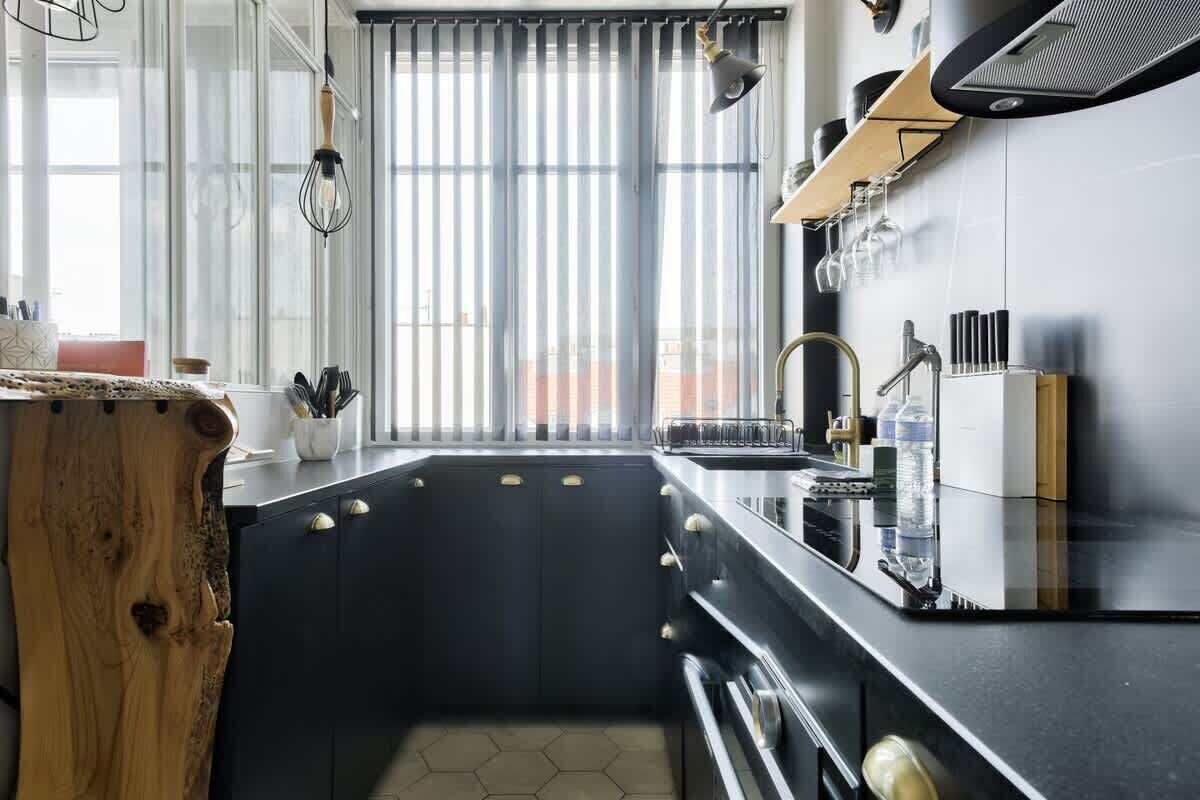 Une cuisine moderne avec un plan de travail noir, des appareils électroménagers en acier inoxydable et une fenêtre équipée de stores verticaux.