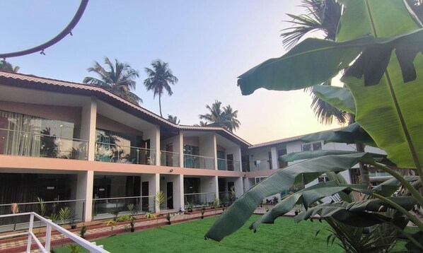 A multi-story building with balconies, a green lawn, and palm trees. - Lexis Inn Beach Resort (Diveagar)