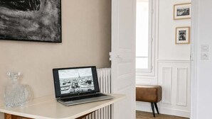 A room with a white radiator, a laptop on a white table, a bench, and a door leading to another room. - Studio Under the Rooftops - Sacre-Coeur View (Paris)