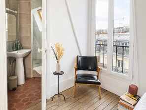 A corner room with a view of the city, a black leather chair, a small table with a vase, and a stack of books. - Studio Under the Rooftops - Sacre-Coeur View (Paris)