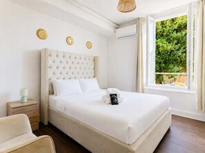 A hotel room with a large bed, a bedside table, a lamp, and a window with a view of greenery. - Séjour idéal climatisé - Paris, CDG - Parking (Le Bourget)