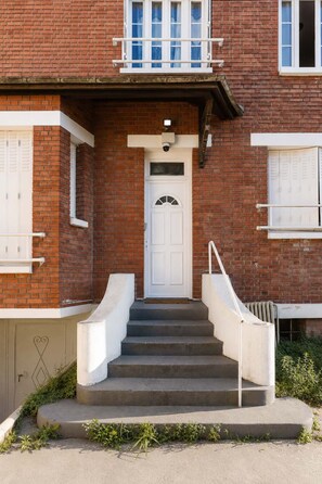 A two-story brick building with a white front door, a small balcony, and a set of concrete steps leading to the entrance. - Séjour idéal climatisé - Paris, CDG - Parking (Le Bourget)