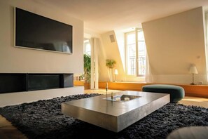 Ein modernes Wohnzimmer mit einem großen Flachbildfernseher an der Wand, einem schlichten Couchtisch und einer gemütlichen Sitzecke mit Sofa und Hocker.