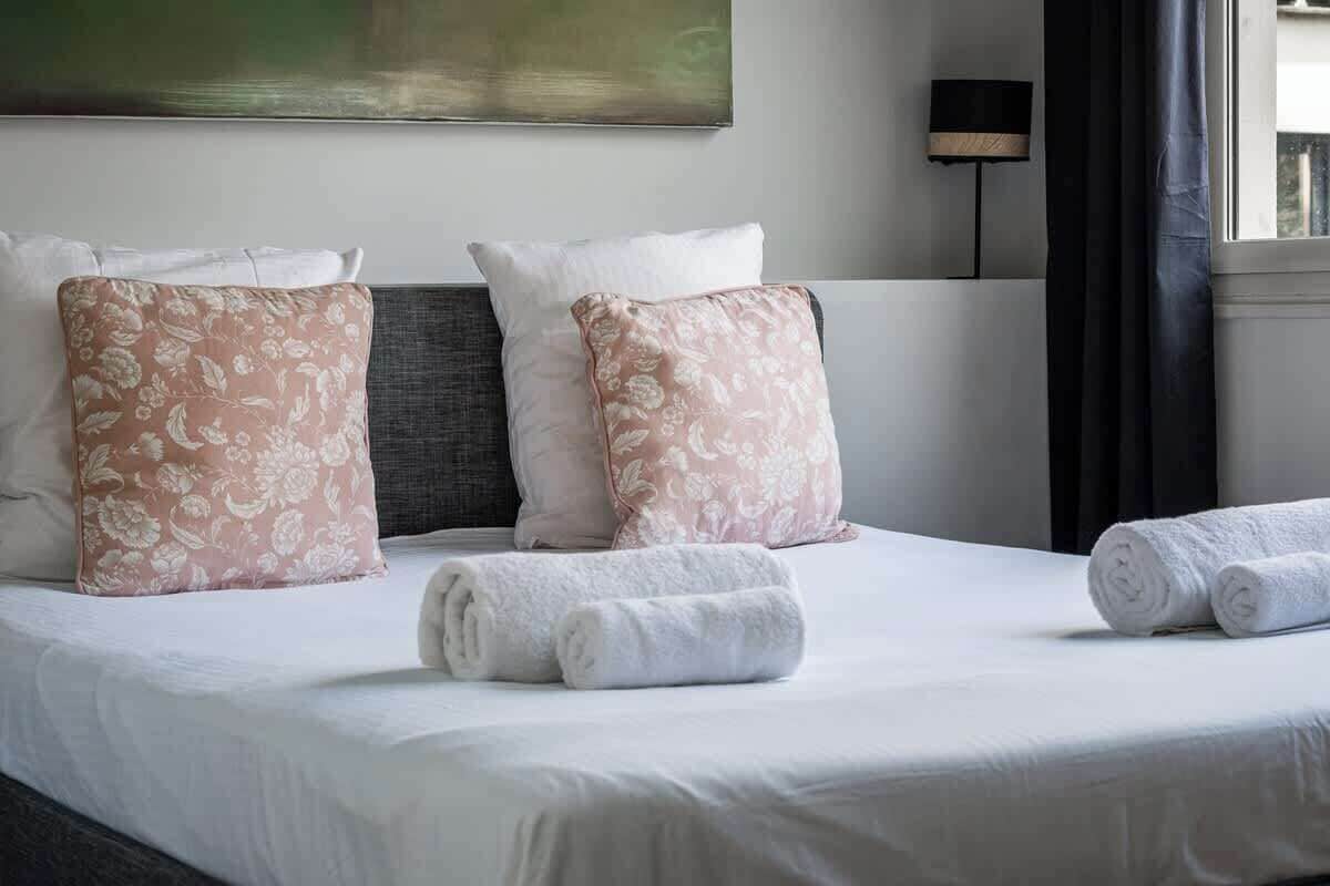 Une chambre d’hôtel avec un lit, deux oreillers, une tête de lit, une lampe fixée au mur et une fenêtre avec des rideaux.