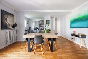 A modern dining area with a wooden table and chairs, a kitchen in the background, and a large abstract painting on the wall. - Spacious fully equipped 1-bedroom - Mobility lease (Paris)