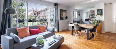 Una sala moderna con un sofá gris, una mesa de centro con un tarro de vidrio, y un comedor con una mesa y sillas de madera.