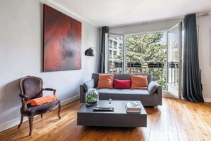 A modern living room with a grey sofa, a wooden chair, a coffee table with a remote and a book, and a large abstract painting on the wall. - Spacious fully equipped 1-bedroom - Mobility lease (Paris)