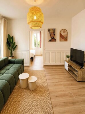 Ein modernes Wohnzimmer mit einem grünen Sofa, zwei runden Beistelltischen, einem Fernseher auf einem Holzgestell und einer großen gewobenen Pendelleuchte.