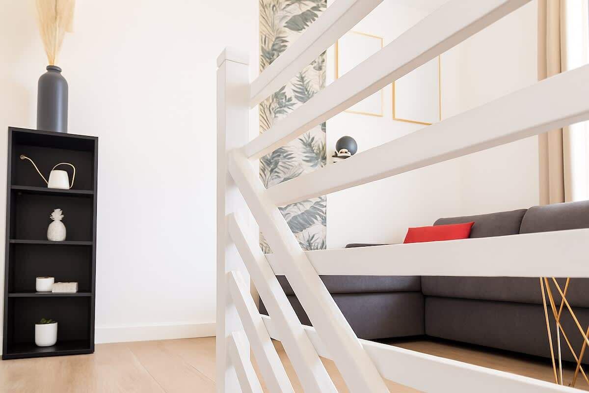 A modern living room with a white staircase, a grey sofa with red cushions, and a black shelving unit with decorative items.