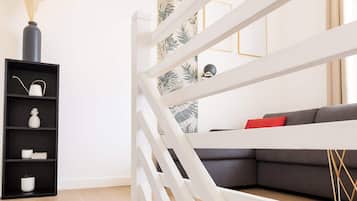 A modern living room with a white staircase, a grey sofa with red cushions, and a black shelving unit with decorative items.