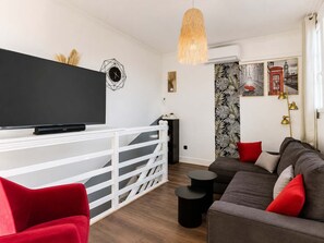 Un saló modern amb un televisor de pantalla plana, un sofà, cadires vermelles, una escala i una paret amb dibuix de fulles tropicals.