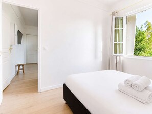 Un dormitori amb un llit, una finestra amb cortines, un tamboret de fusta i una porta que dona a una altra habitació.