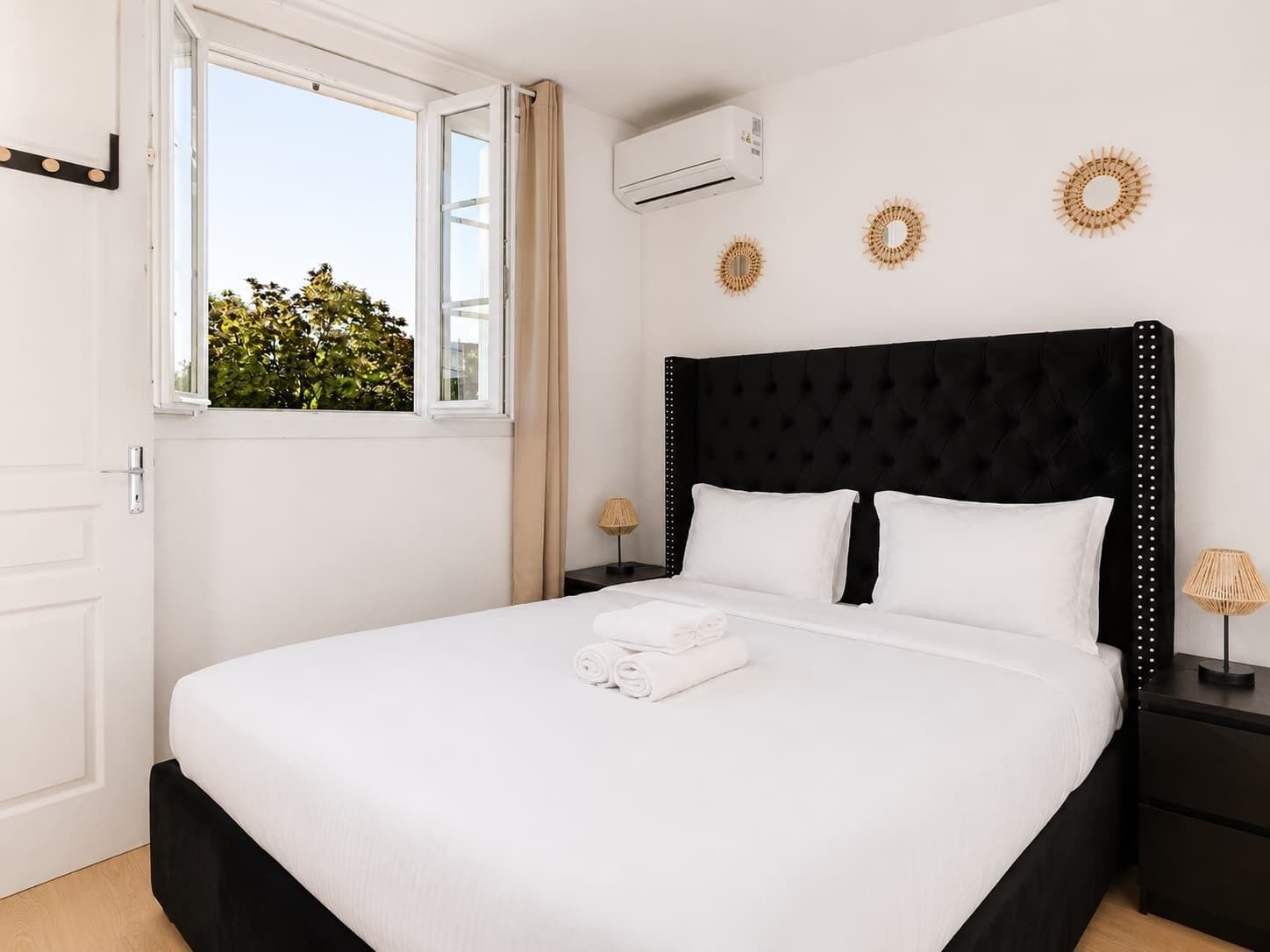 A bedroom with a black headboard, white bedding, and a window with a view of greenery.