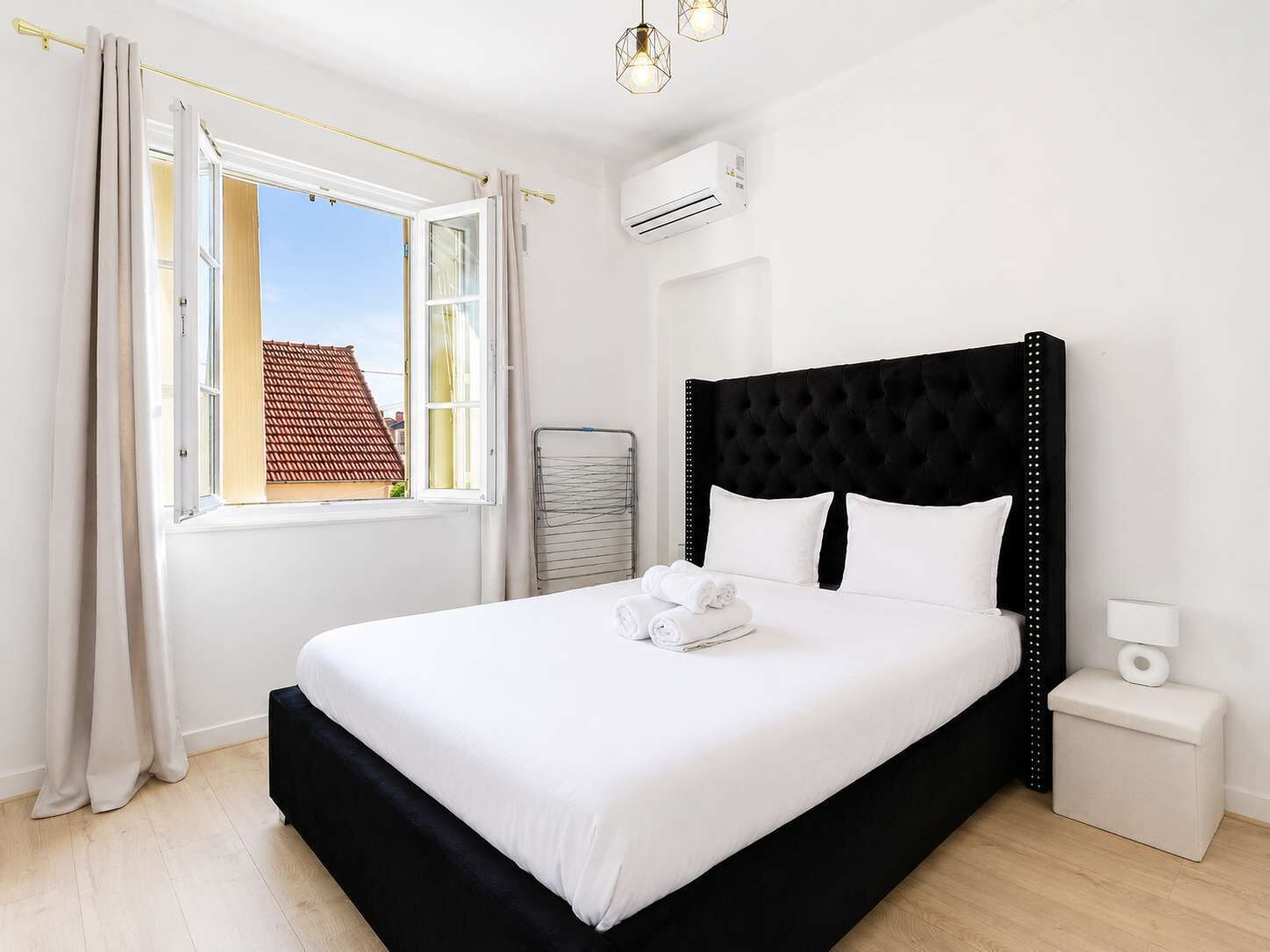 A hotel room with a bed, a nightstand, an air conditioning unit, a window with curtains, and a view of a building outside.