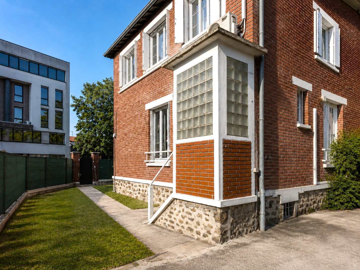 Een tweekamerig bakstenen gebouw met een stenen plint, grote ramen en een kleine voortuin.