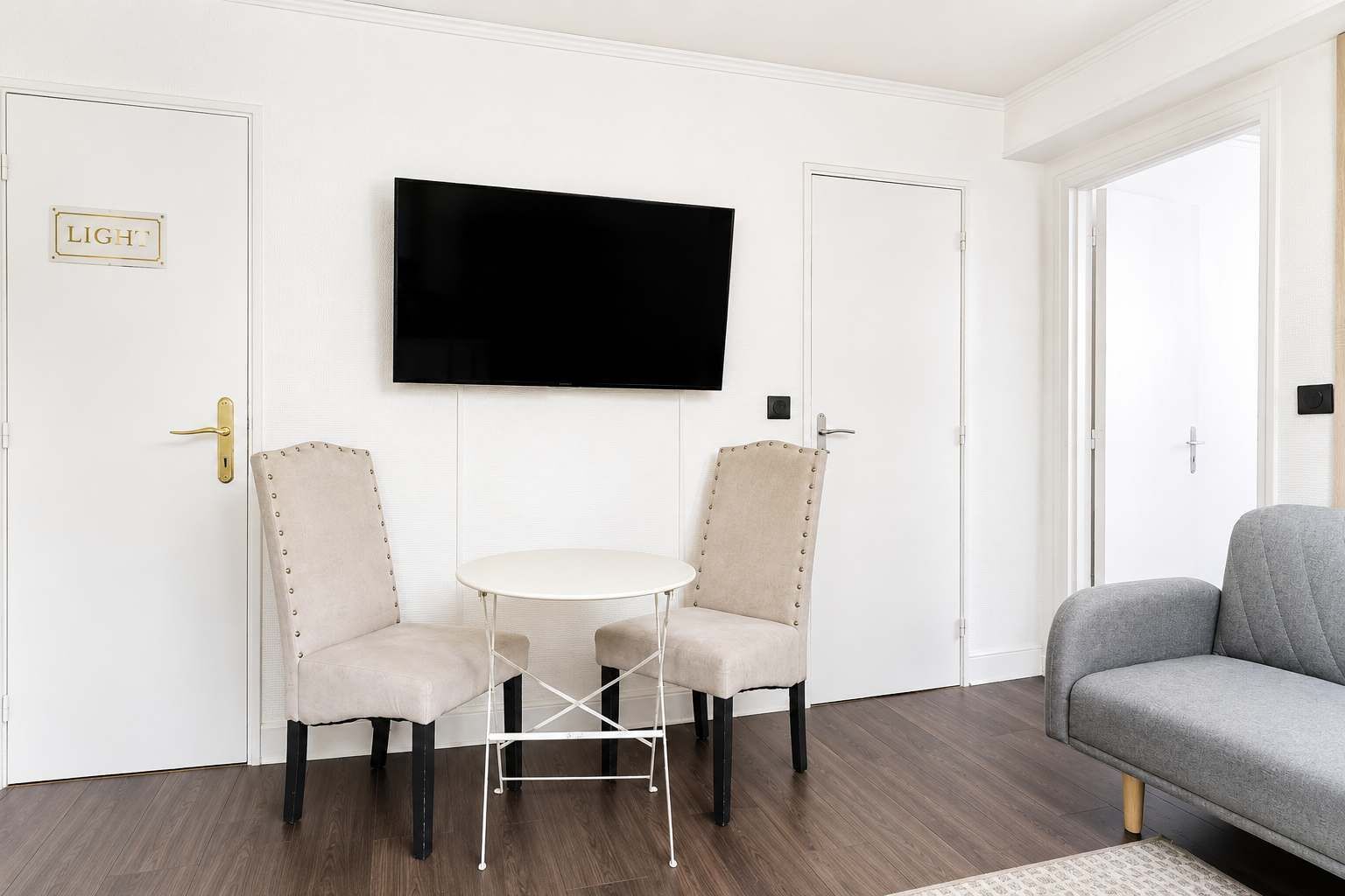 Een kamer met een wandgemonteerde flatscreen televisie, twee gestoffeerde stoelen met metalen poten, een klein rond tafeltje en een grijze bank.