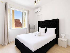 A bedroom with a large bed, a window with curtains, an air conditioning unit, and a bedside table with a lamp. - Havre cosy climatisé - Parking (Le Bourget)