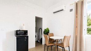 Ett kompakt bostadsutrymme med ett litet kök, en matplats med ett runt bord och stolar samt ett fönster med gardiner.