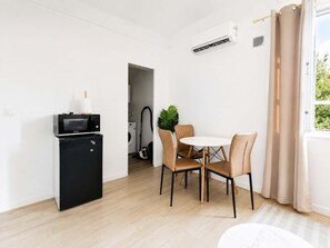 A compact living space with a kitchenette, a dining area with a round table and chairs, and a window with curtains. - Havre cosy climatisé - Parking (Le Bourget)