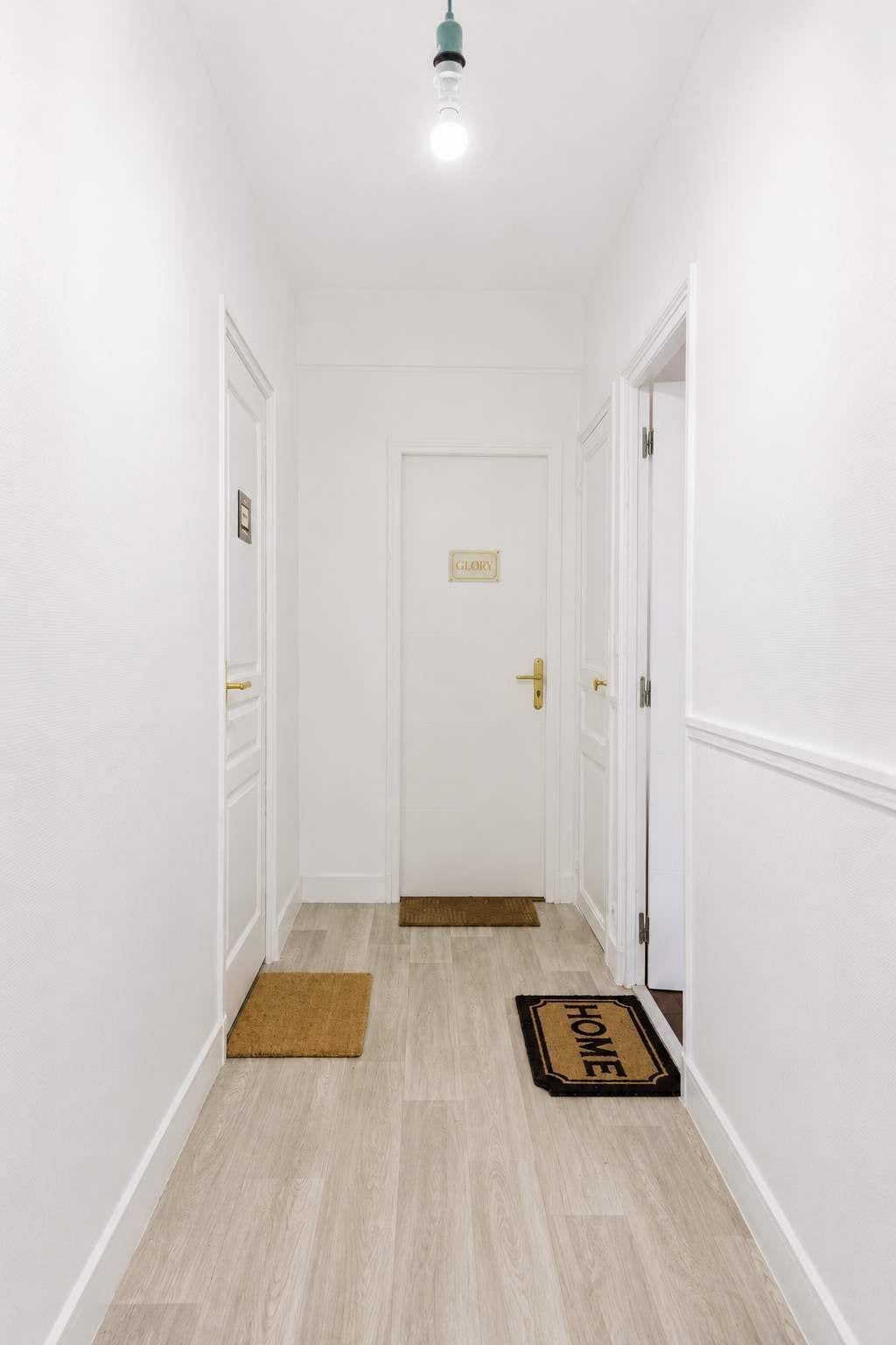 A hallway with white walls, a wooden floor, a door with a 'GLORY' sign, and a doormat with 'HOME' on it.