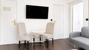 A room with a flat-screen TV mounted on the wall, two upholstered chairs with metal legs, a small round table, and a grey sofa. - Appartement lumineux climatisé - Parking (Le Bourget)