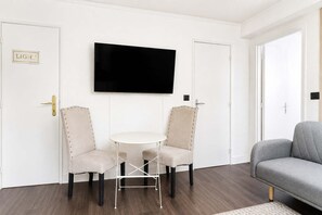 Ein Zimmer mit einem Wand-Fernseher, zwei Polsterstühlen mit Metallbeinen, einem kleinen runden Tisch und einem grauen Sofa.