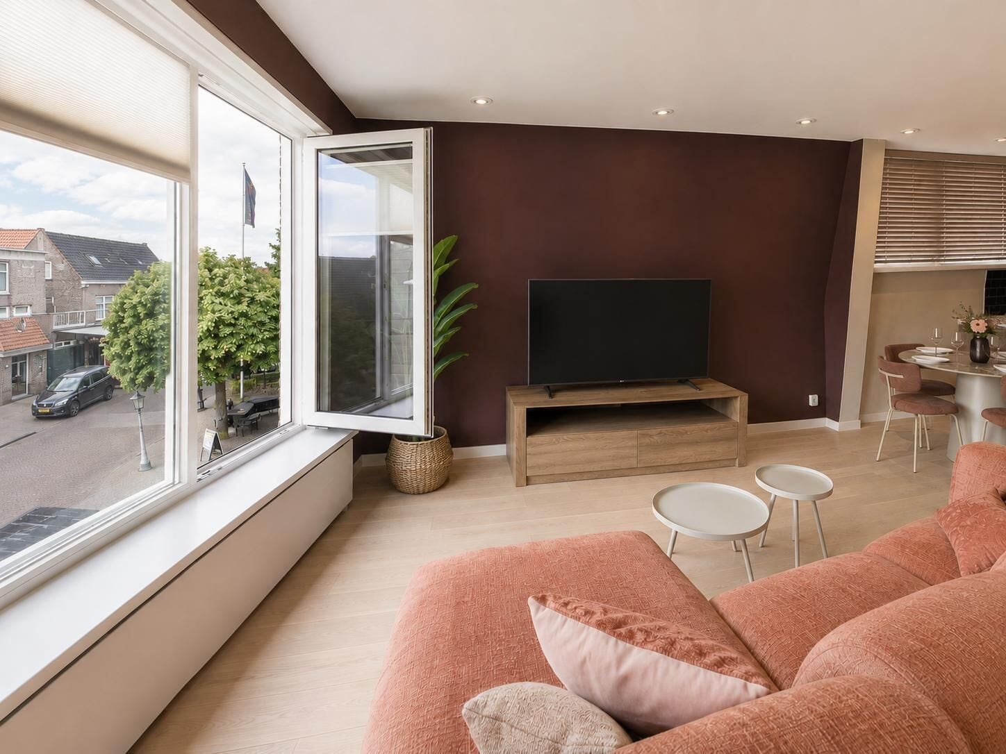 Een moderne woonkamer met een groot raam, een flatscreen televisie, een houten tv-meubel, een roze bank en een eethoek met een tafel en stoelen.