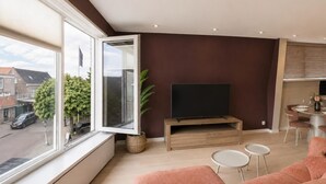 A modern living room with a large window, a flat-screen TV, a wooden entertainment unit, a pink sofa, and a dining area with a table and chairs. - Historic Stay in Medemblik | Tesstays (Medemblik)