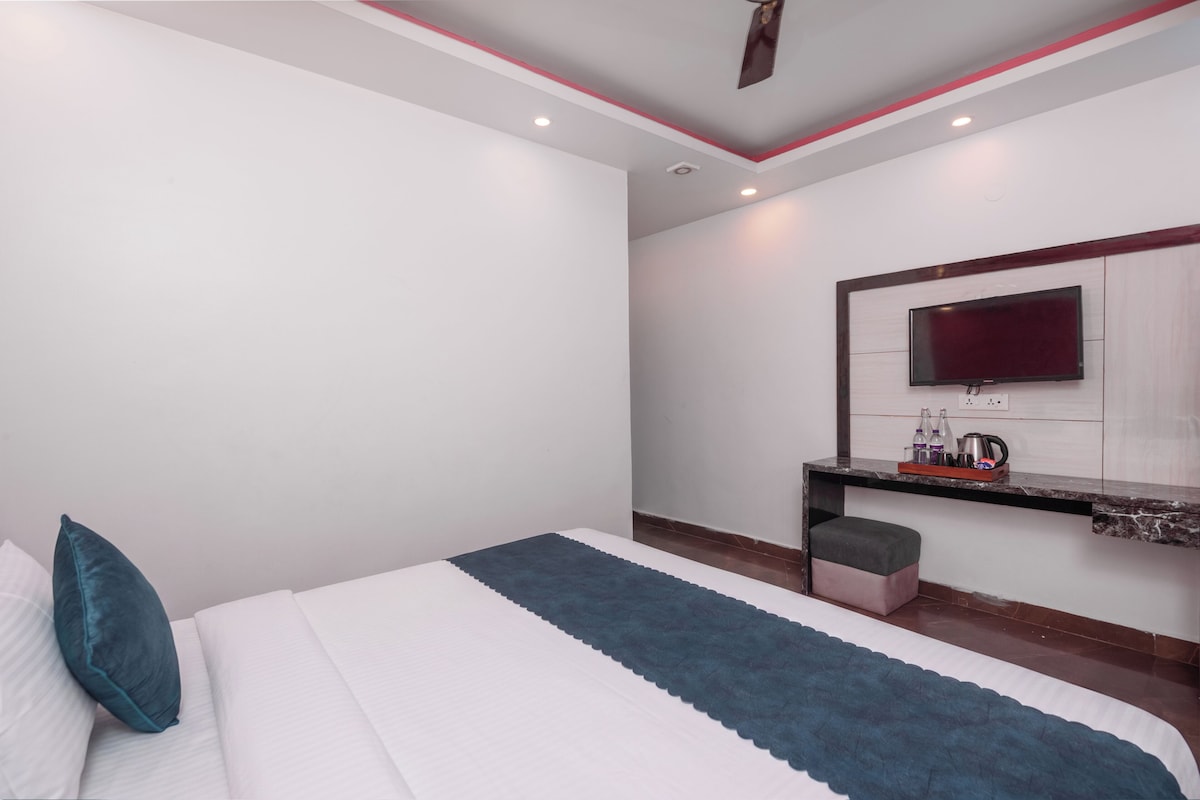 Habitación de hotel con cama, televisión montada en la pared, una mesita con una tetera y botellas, y un ventilador de techo.