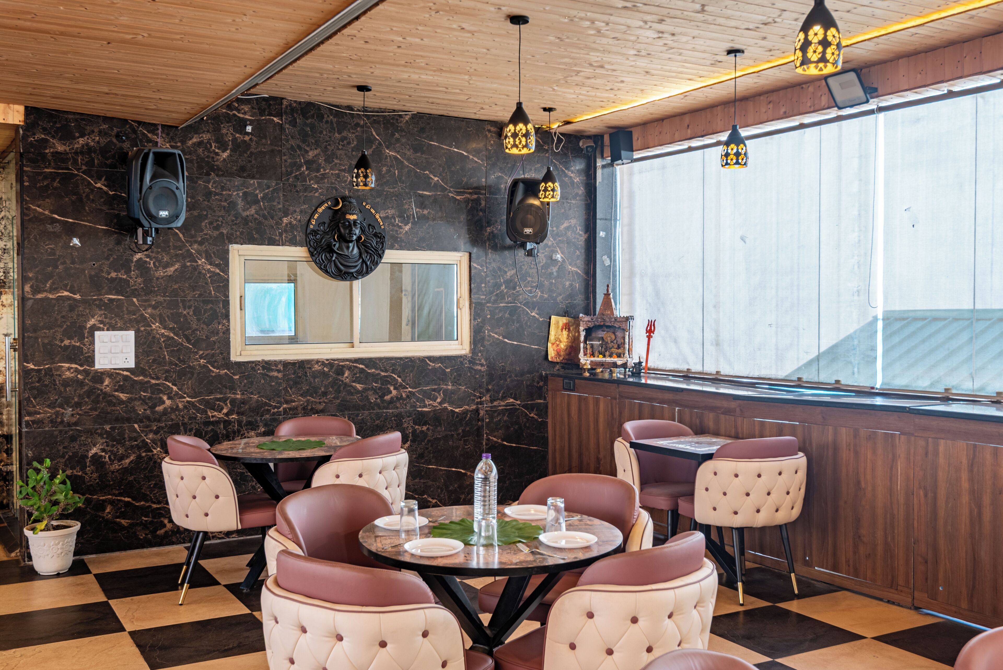 A restaurant interior with marble walls, wooden ceiling, and black and white checkered flooring. There are round tables with chairs, and pendant lights hanging from the ceiling.