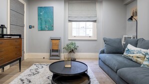 Una sala de estar con un sofá gris, una mesa de centro, un aparador de madera, un cuadro, una ventana con persianas y un radiador.