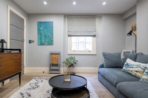 A living room with a grey sofa, a coffee table, a wooden dresser, a painting, a window with blinds, and a radiator. - Chic 1BDR City Retreat by Regents Park (Greater London)