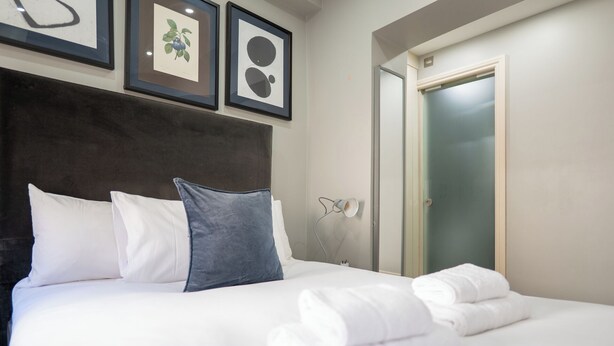 Una cama bien hecha con almohadas blancas y azules, un cabecero y una lámpara de pared.
