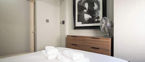 A hotel room with a bed, a framed abstract painting, a wooden dresser, and a standing fan.