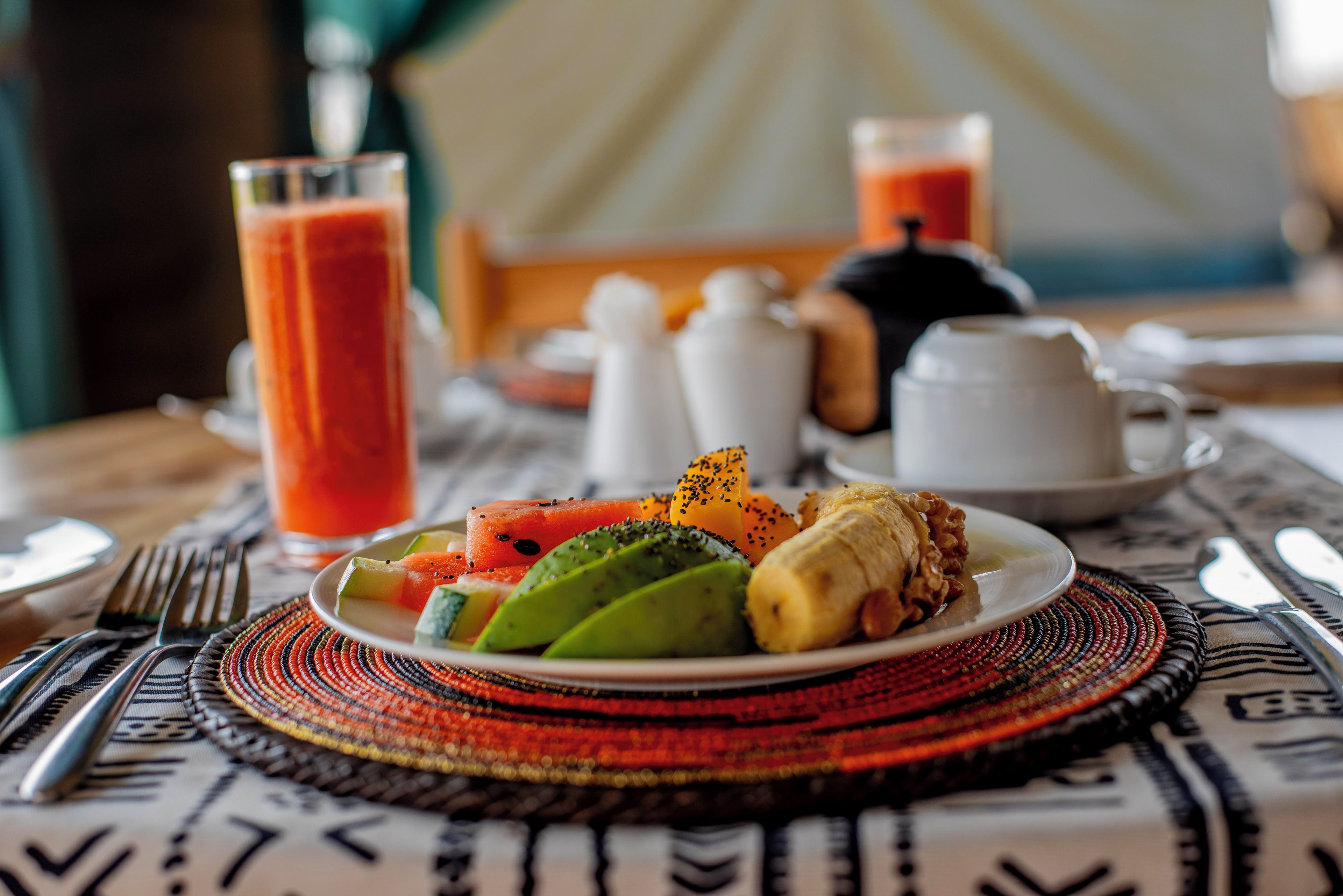 Disposição para o café da manhã com uma travessa de frutas, um copo de suco e uma chaleira sobre uma mesa com jogo americano estampado.