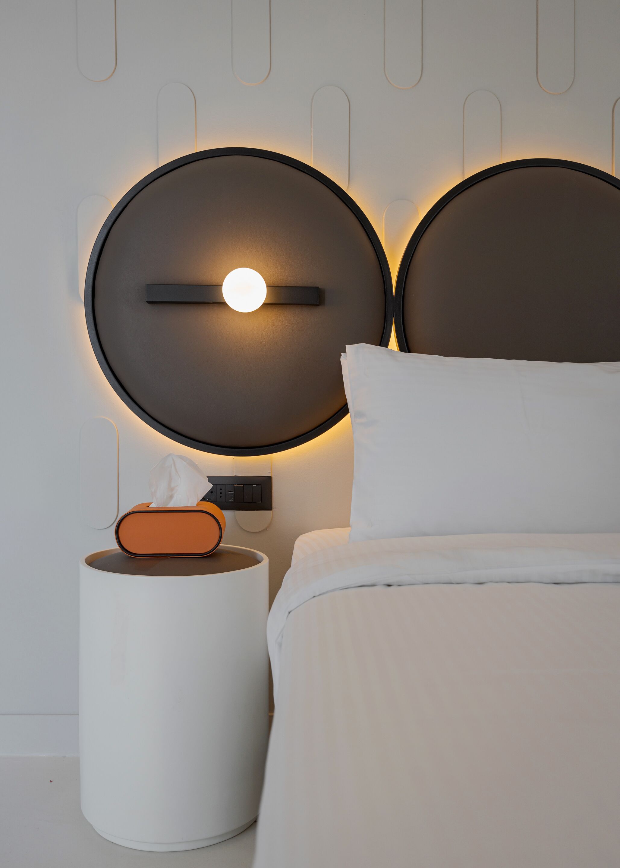 A modern bedroom with a bed, a bedside table with a tissue box, and a wall-mounted light fixture.