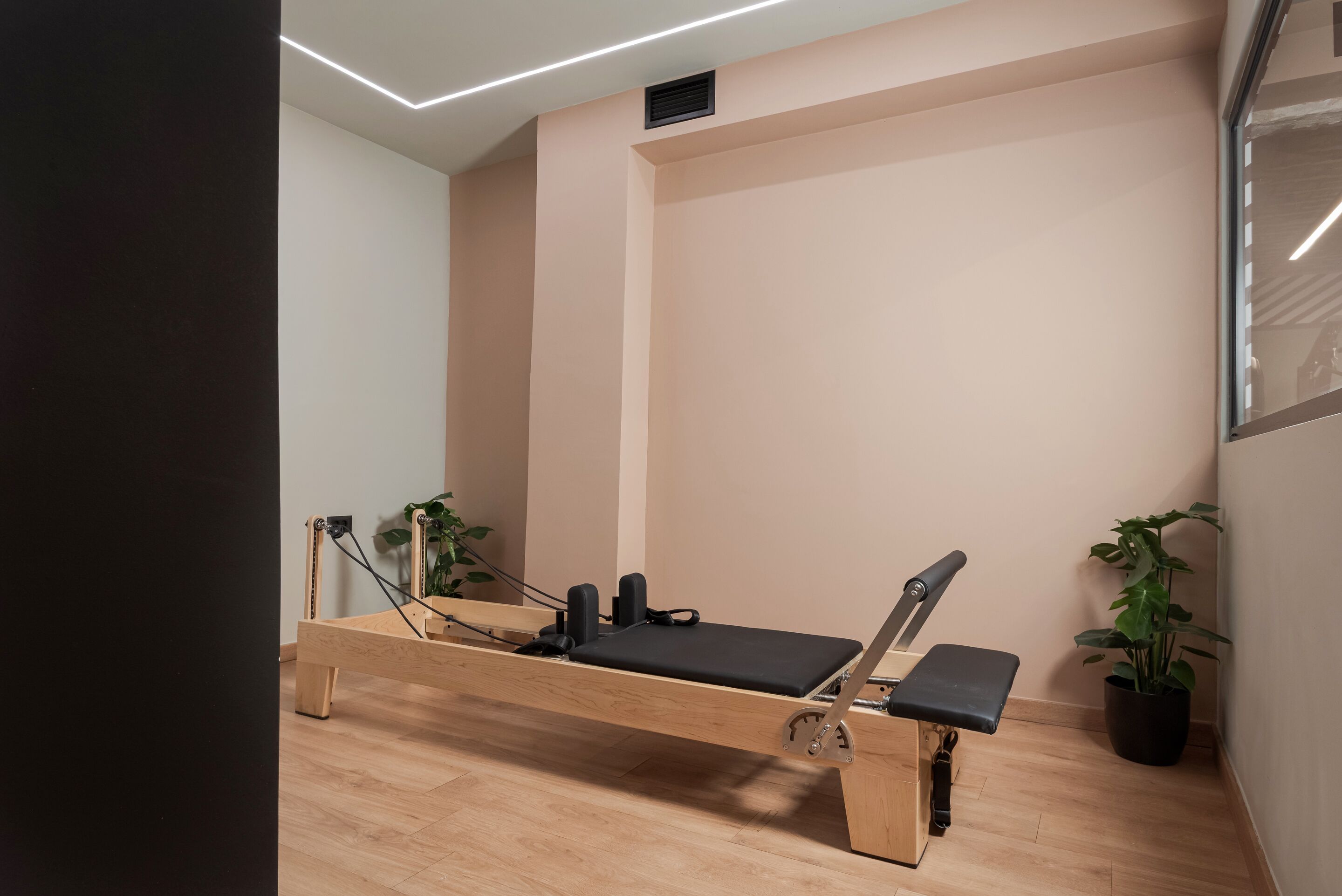 En Pilates-reformer i et rum med trægulv, et spejl på væggen og to potteplanter.